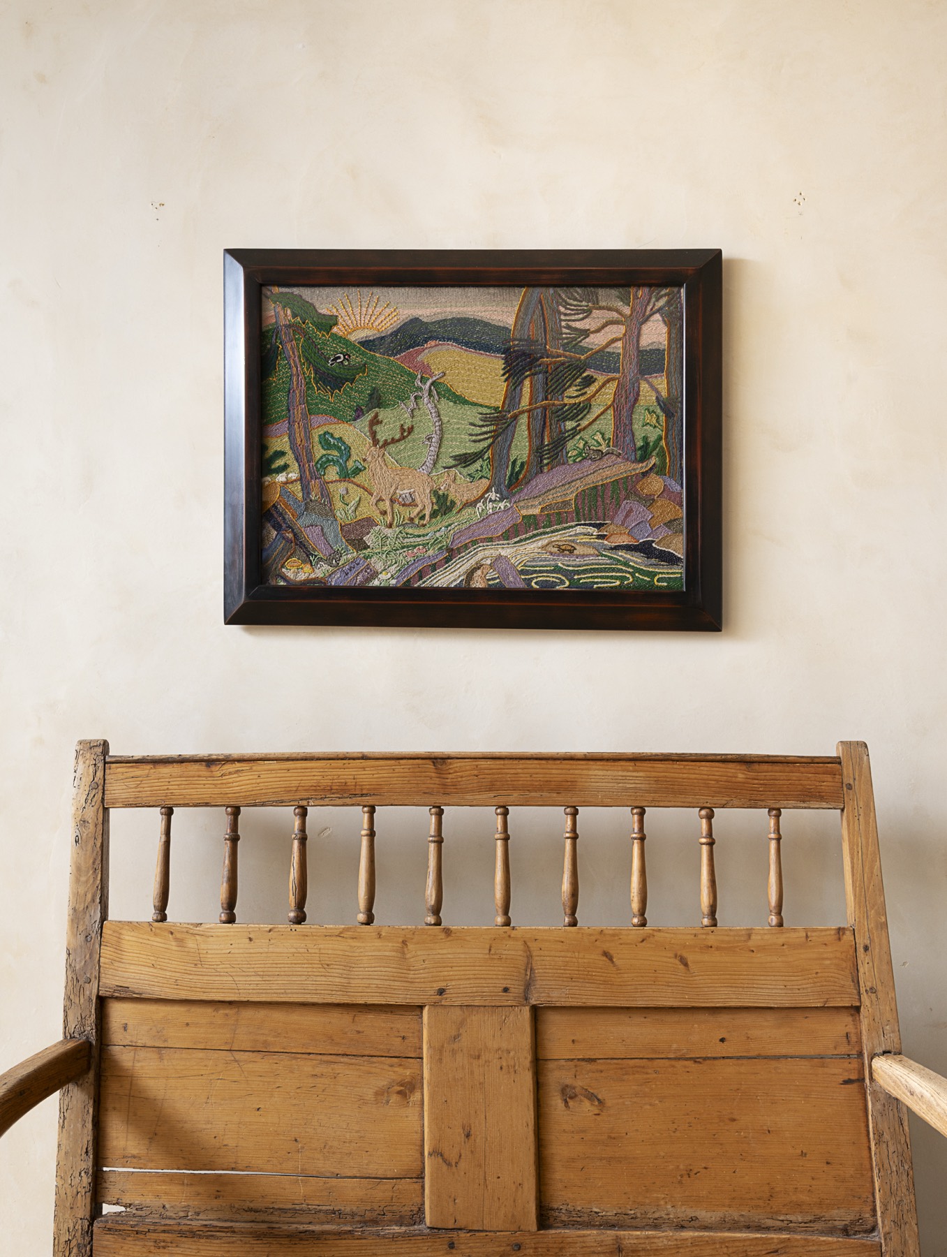 Folk embroidered landscape in a dark frame above a pine bench