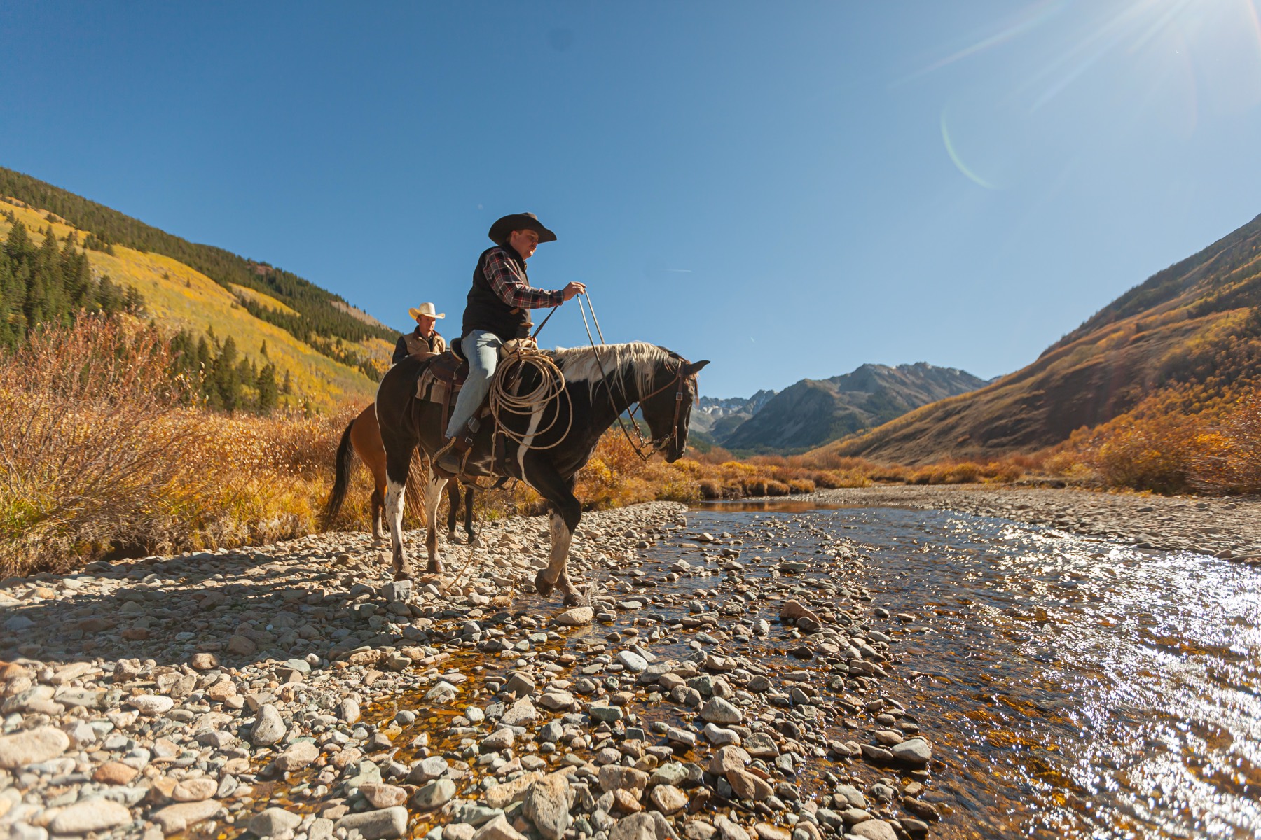 Two cowboys on horseback crossing a shallow river beneath autumn hillsides