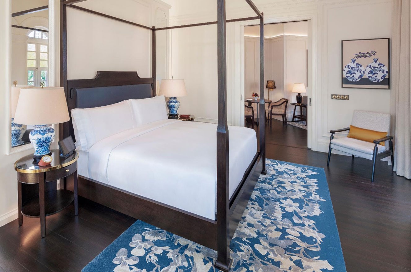 A Raffles Singapore guest room: dark four-poster bed, blue-and-white porcelain lamps, a blue floral rug, and a framed painting of koi vases on the far wall