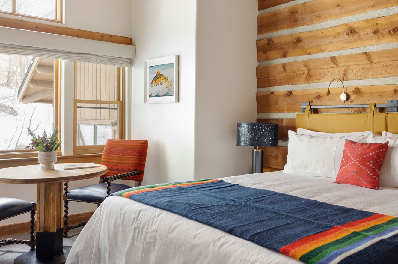 Sundance model guest room: a framed trout watercolor beside the window, log wall, mustard headboard, and a striped wool blanket on the bed