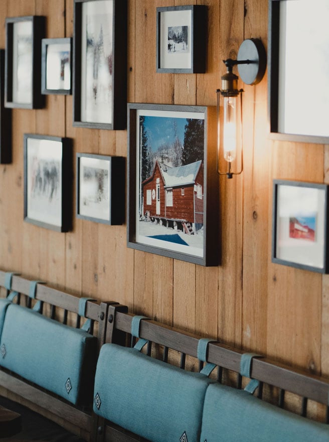 Gallery wall of framed vintage ski photographs on cedar planking above teal banquette seating at The Lookout, with a red cabin in snow at center